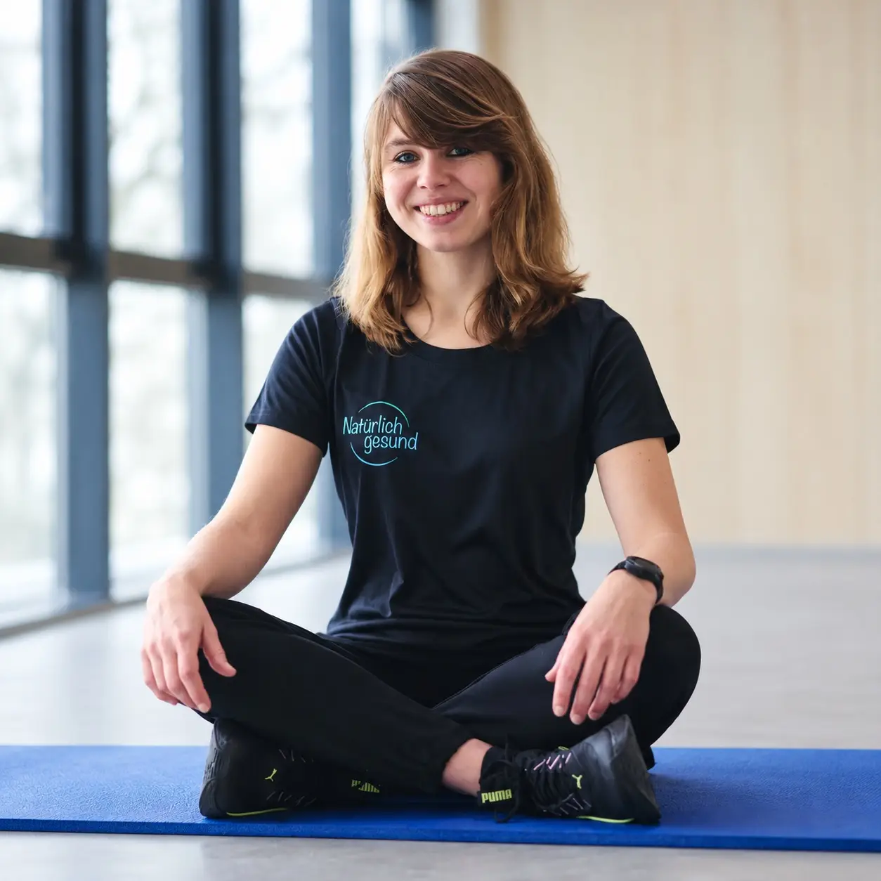 Tahnee Boderius sitzt im Natürlich-gesund-Shirt im Schneidersitz auf einer Gymnastikmatte und lächelt offen in die Kamera